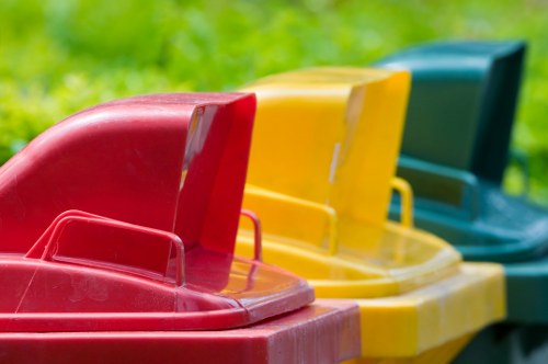 The image shows a row of three large plastic wheelie bins positioned outdoors on a grassy area, possibly within a garden or driveway. The bins are arranged side by side, with the one in the foreground being red, featuring a sturdy handle and a slightly textured, glossy surface. Behind it, a yellow bin with similar design elements is visible, followed by a green bin at the back. The bins are placed on a lush green lawn or garden space, with a blurred background suggesting natural surroundings. The lighting appears natural, highlighting the vibrant colours and smooth finish of each bin. This scene is consistent with a waste or rubbish removal service, such as those offered by Garden Clearance Brimsdown, specialising in outdoor waste collection and clearance services in the local area near Brimsdown postcode regions. The image emphasizes the type of waste containers used for garden or outdoor refuse collection, relevant to rubbish removal efforts for residential or commercial clients.