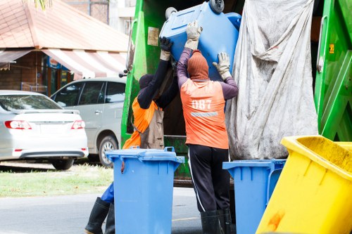 Two waste management workers wearing high-visibility orange vests and dark clothing are positioned in front of a large green waste collection vehicle on a residential street, likely in Brimsdown. They are handling a large blue plastic bin, with one worker reaching up to lift or secure a blue cylindrical waste container, which appears to be made of durable plastic. The worker on the right, wearing a head covering, is guiding or steadying the bin while the worker on the left supports it from below. Nearby, two more blue bins and a yellow waste wheelie bin are visible on the street, indicating ongoing rubbish collection and outdoor waste removal activities. In the background, there are parked cars, a tree-lined pavement, and residential buildings with brick facades and awnings, suggesting a suburban setting. The natural daylight and the urban environment align with typical rubbish collection operations, showcasing the professionalism of Garden Clearance Brimsdown's outdoor waste removal services servicing local communities.