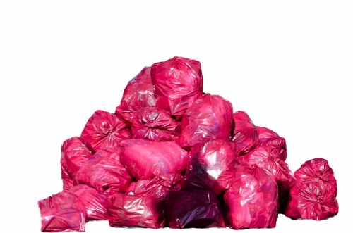 A large pile of bright pink plastic rubbish bags, stacked unevenly with some bags leaning against others, occupying the centre of the image. The bags are filled with waste material, creating a bulky appearance with visible creases and shiny textured surfaces. The background is plain white, providing no environmental context or additional objects. The bags are uniformly tied at the tops, with some slightly bulging at the sides and showing irregular shapes. This scene relates to waste collection or rubbish removal services, such as those offered by Garden Clearance Brimsdown, with the focus on a substantial quantity of outdoor waste ready for disposal or collection.