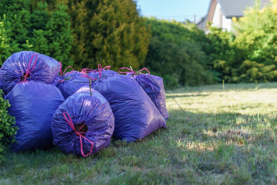 A black wheeled rubbish bin positioned outdoors on a grassy area, filled with a mix of garden waste including dry, beige grass or reeds, green leafy branches, and other organic debris. The bin is made