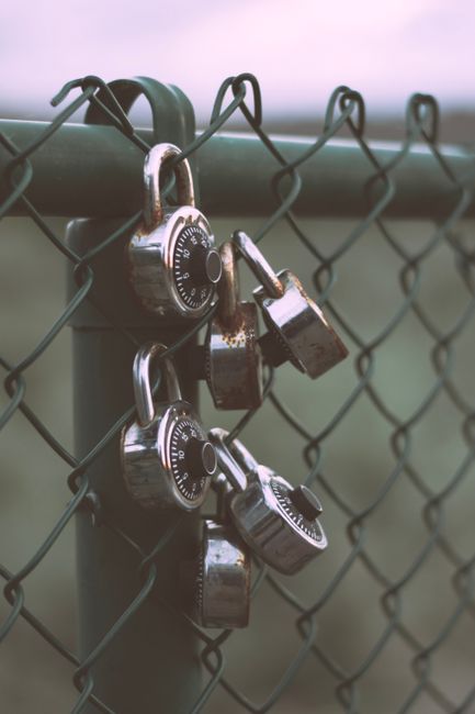 Close-up of a metal chain-link fence with three combination padlocks attached to the corner where two fence panels meet. The padlocks are silver with black dials, showing different numbers, some sligh